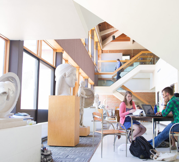Two people study at a table in a bright, modern space with large windows, sculptures, and visible stairs; another person is seen on the upper level.