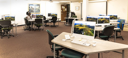 A computer lab with several desks and iMac computers, mostly empty except for one person working at a computer.