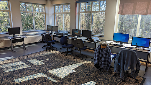 A sunlit computer lab with several desks, chairs, desktop computers, and a patterned rug. Large windows show trees outside. Some chairs have jackets draped over them.