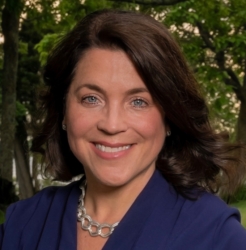 A woman with shoulder-length brown hair, wearing a navy blazer and a silver necklace, smiles outdoors with green trees in the background.