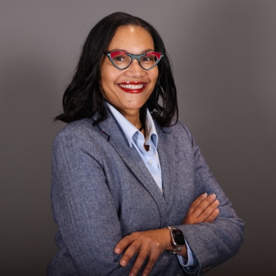 A person with long black hair and red lipstick is smiling. They wear red and black glasses and a blue blazer.