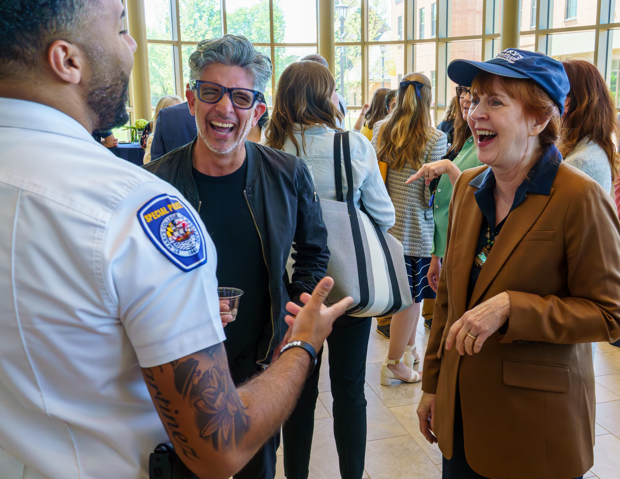 A security officer talks and laughs with a man in sunglasses and a woman in a brown blazer at an indoor social event. Other people mingle in the background.
