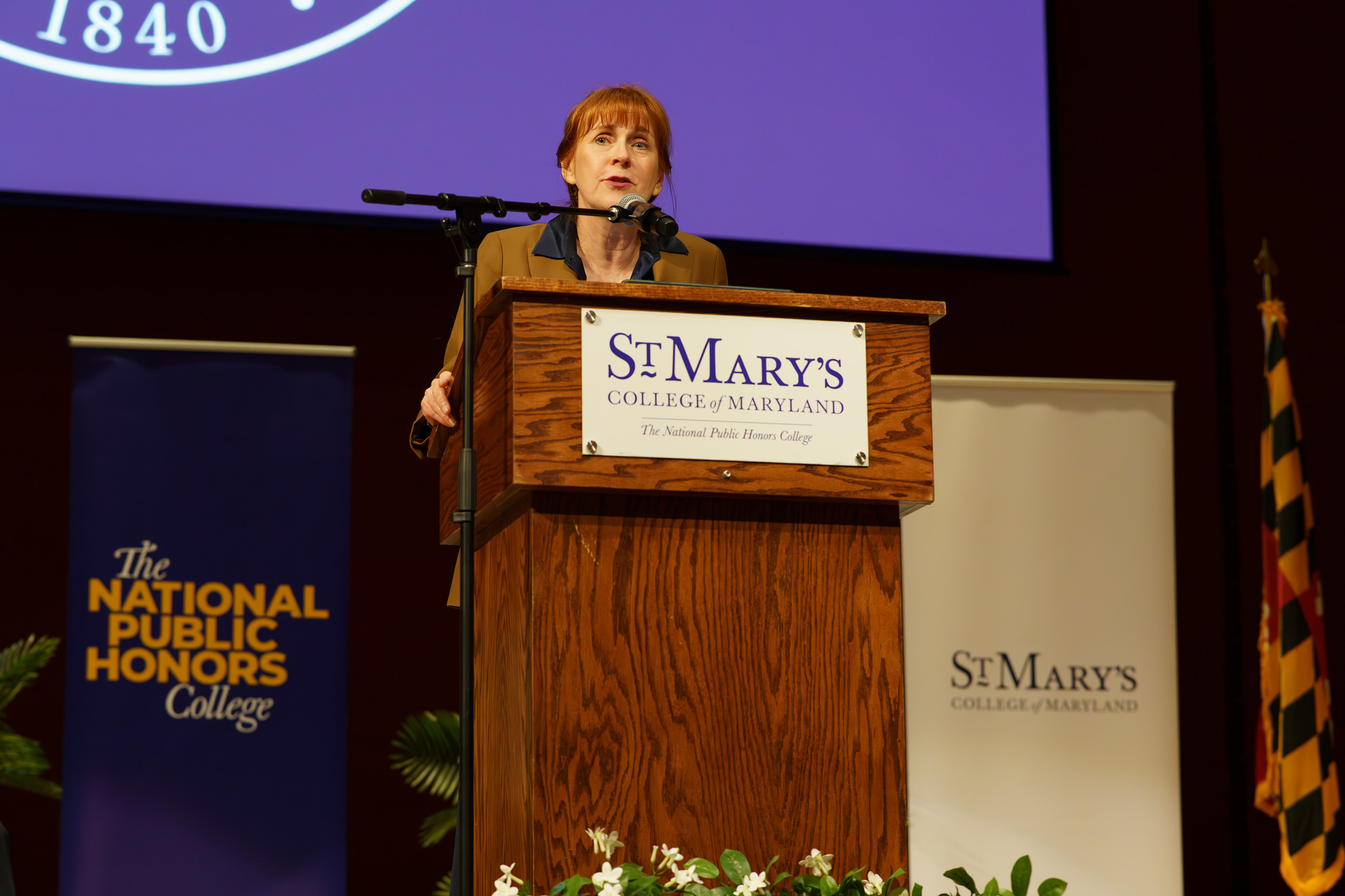 A woman stands at a podium labeled "St. Mary's College of Maryland," delivering a speech at an indoor event. Banners for St. Mary’s College and National Public Honors College are visible.