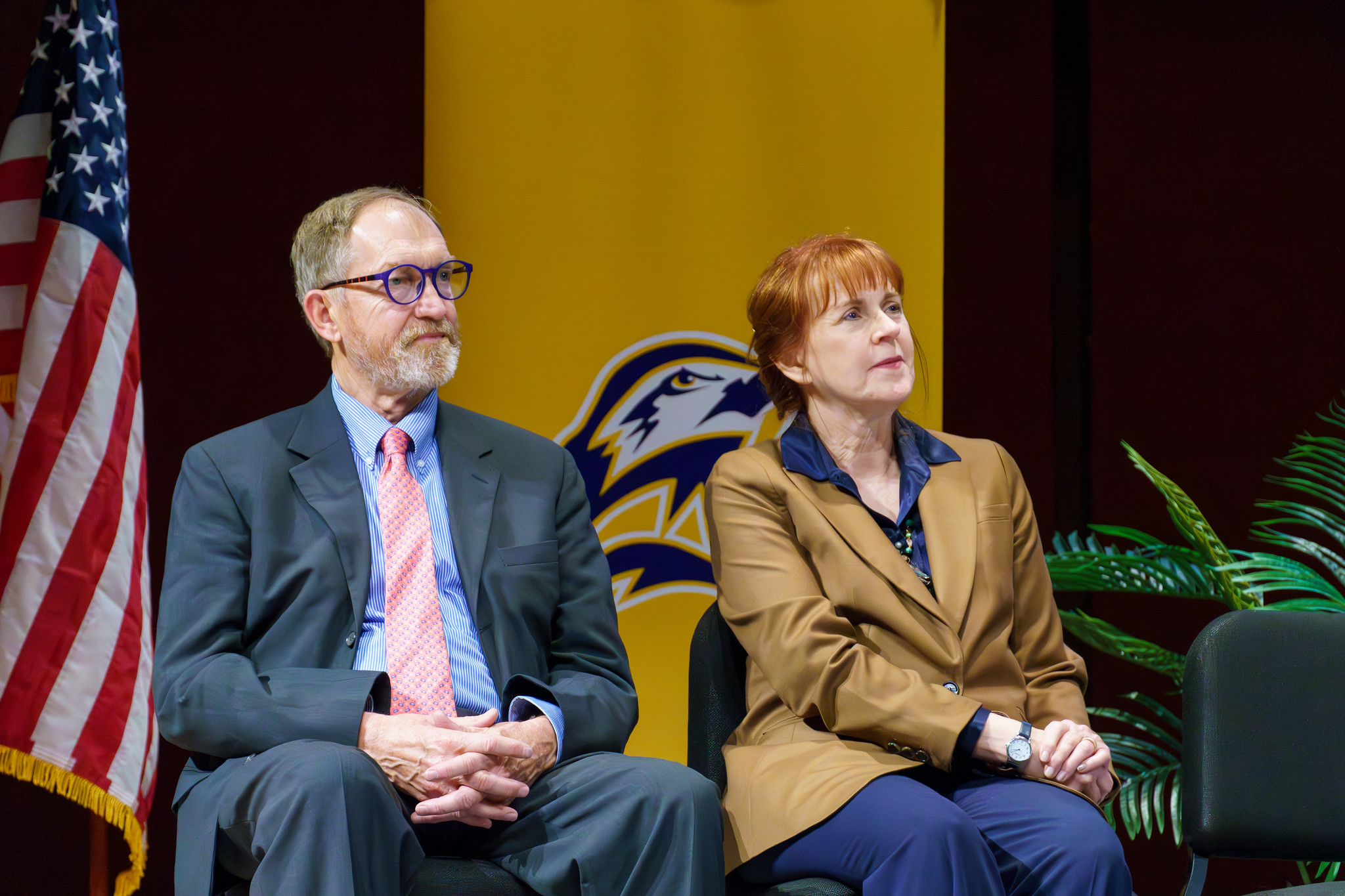 Two adults in business attire sit on stage in front of a yellow banner with an eagle logo and a U.S. flag, appearing attentive during an event.
