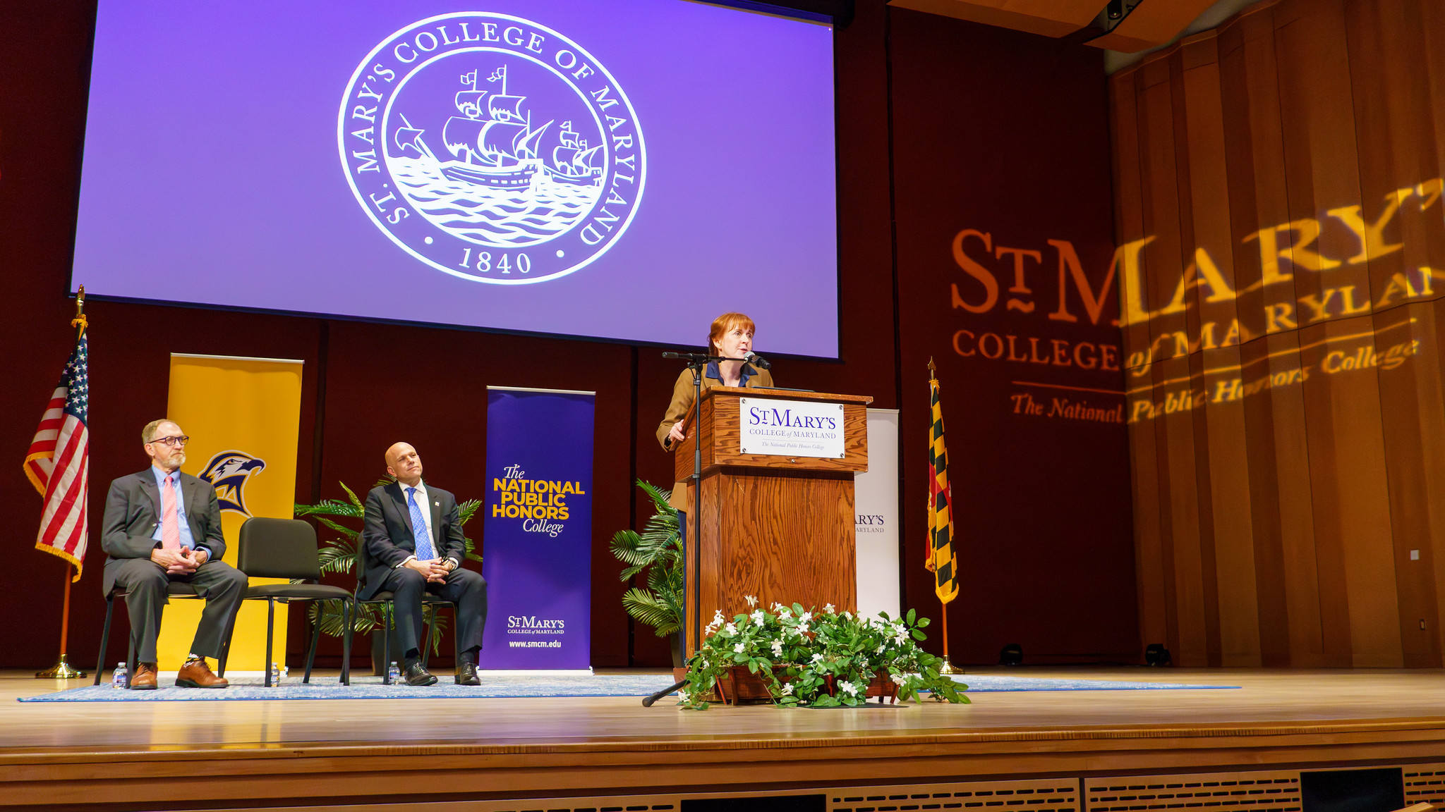 A person speaks at a podium on a stage at St. Mary’s College of Maryland, with two people seated nearby and banners and flags in the background.