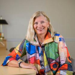 A woman with blonde hair wearing a colorful patterned jacket sits at a table, smiling, with a vase of flowers and a lamp in the background.