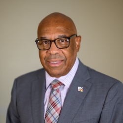 A man wearing glasses, a gray suit, and a plaid tie stands in front of a plain background, smiling slightly at the camera.