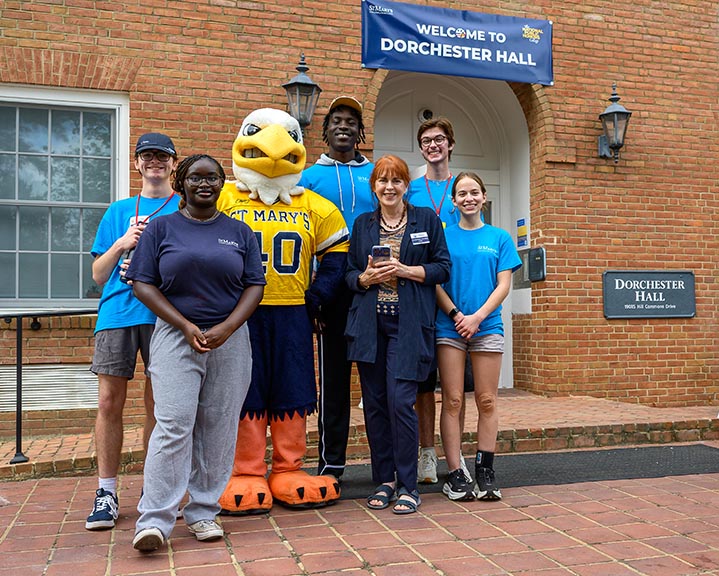 A group of six people, including a person in a bird mascot costume, stand smiling in front of Dorchester Hall with a "Welcome to Dorchester Hall" sign above the door.