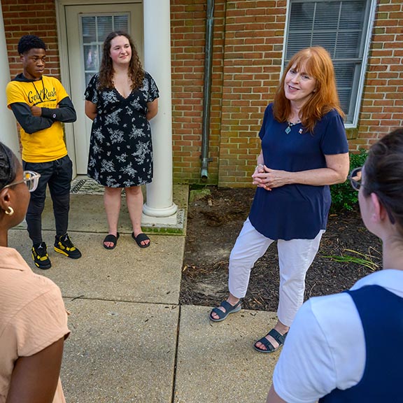 A woman speaks to a group of four people standing in a semicircle outside a brick building.