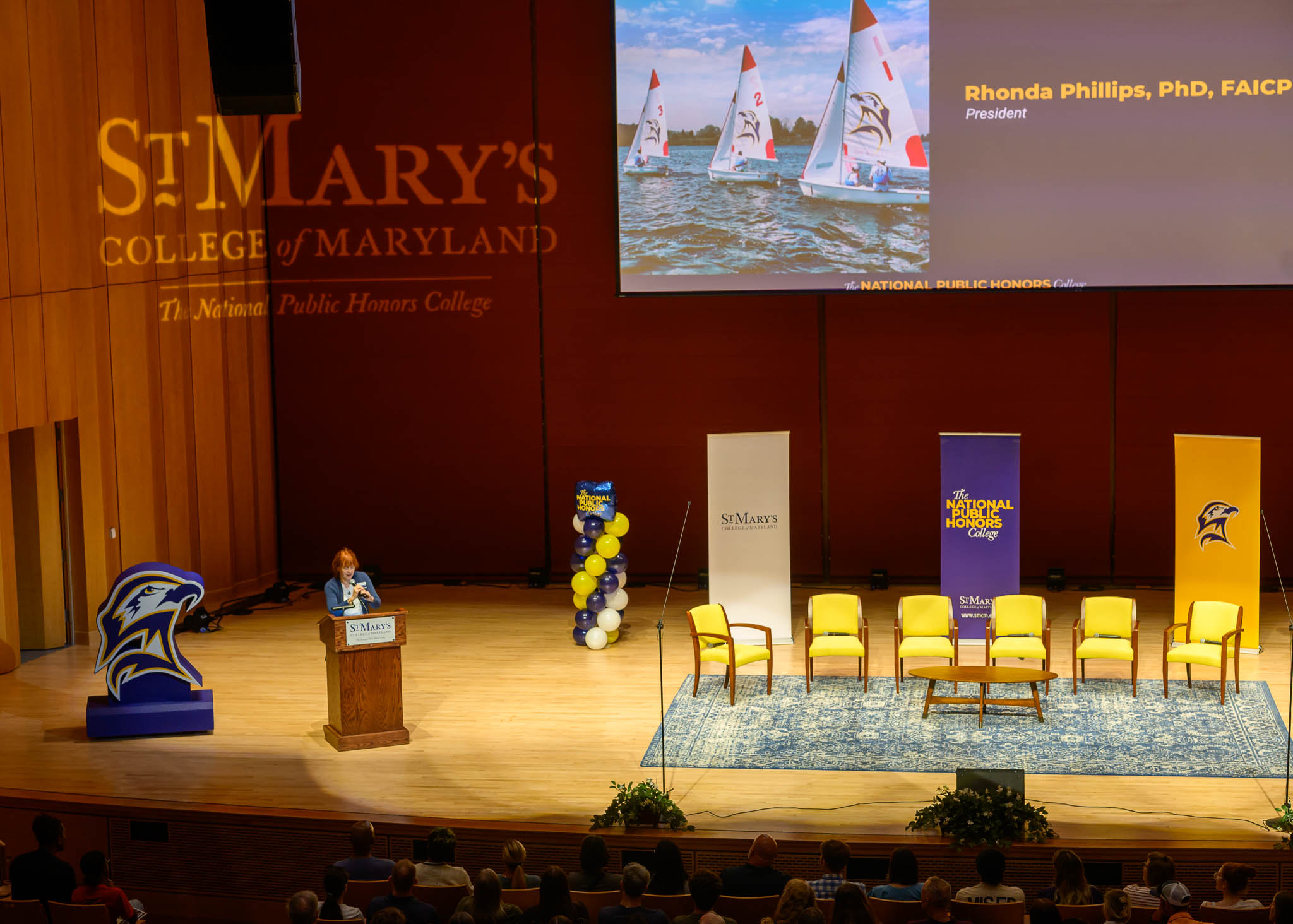 A speaker stands at a podium on a stage with yellow chairs, balloons, and banners at St. Mary's College of Maryland event. A screen displays text and an image of sailboats.