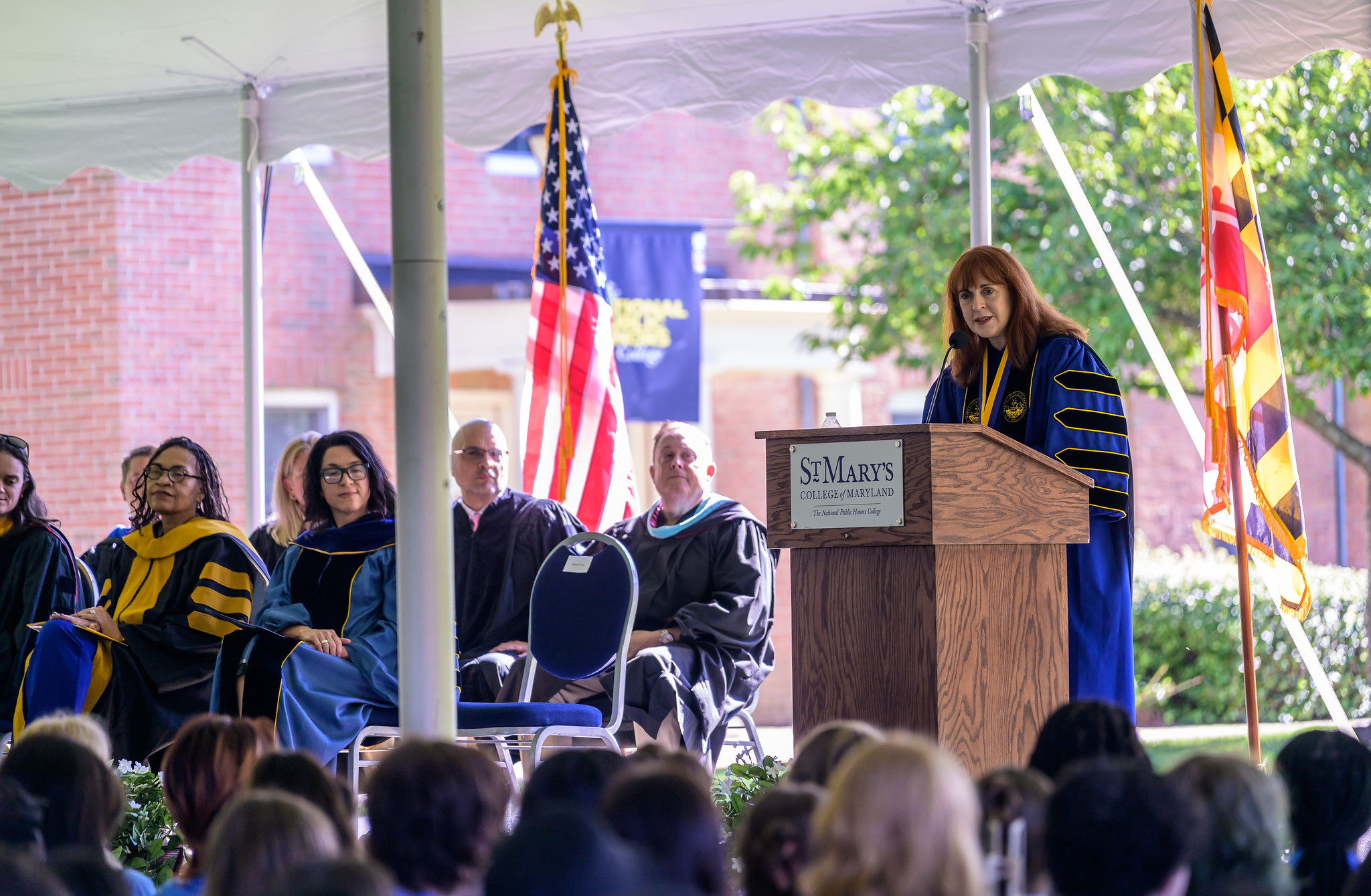 A woman in academic regalia speaks at a podium during an outdoor ceremony, with faculty seated beside her and flags displayed in the background.