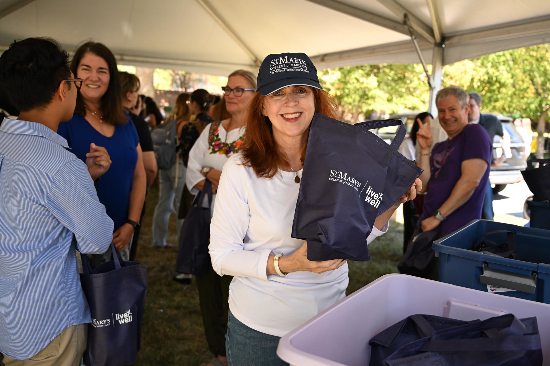 A woman wearing a St. Mark’s hat smiles and holds a navy blue tote bag at an outdoor event under a tent, with other people and bins visible nearby.