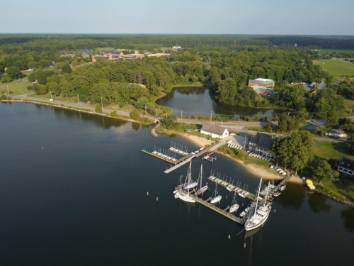 Aerial view of a marina with several docked sailboats, surrounded by trees, water, and nearby buildings on a clear day.