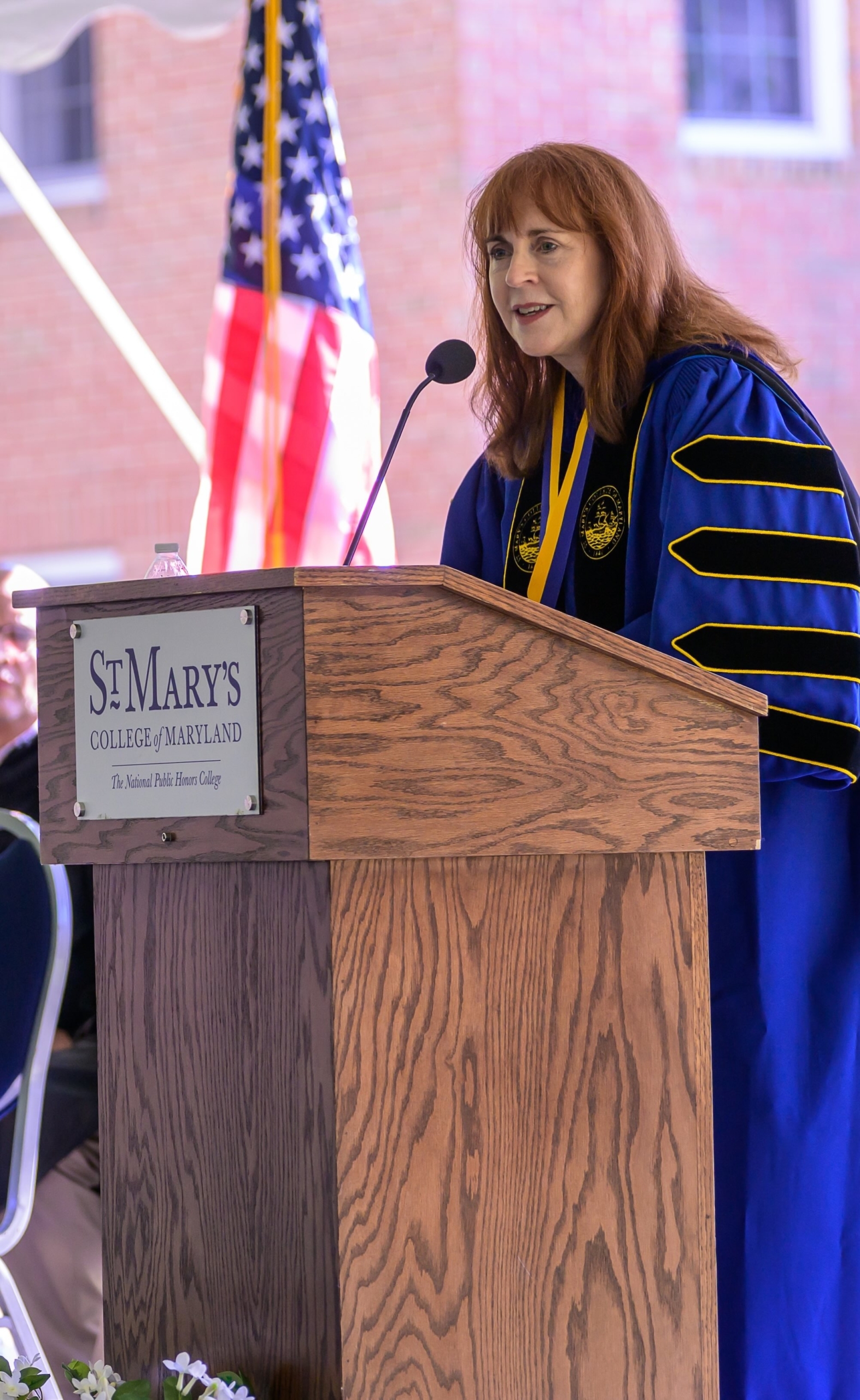 A woman in academic regalia speaks at a podium labeled "St. Mary's College of Maryland" with an American flag in the background.
