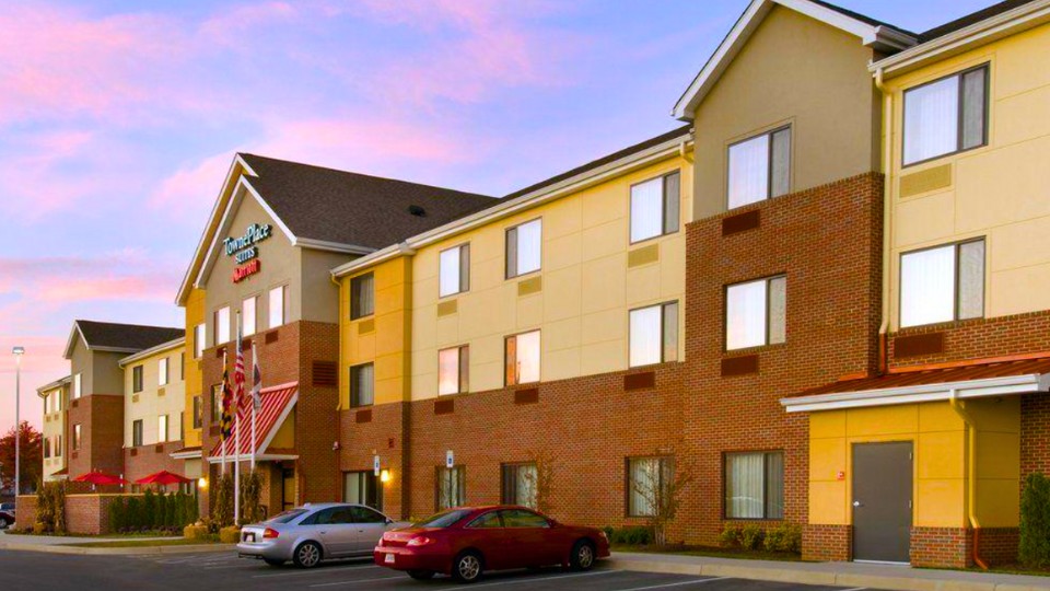 Three-story hotel building with brown and yellow exterior, numerous windows, parked cars in front, and American flags by the entrance at sunset.