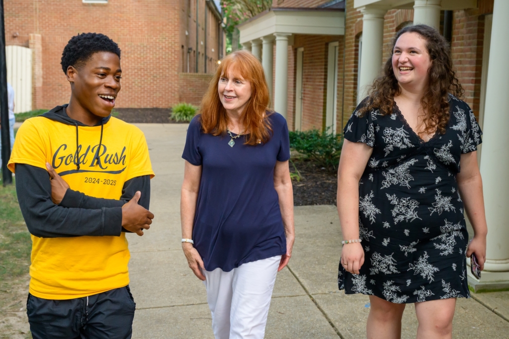 Three people, two women and one man, walk together outside on a sidewalk, smiling and talking, with brick buildings and greenery in the background.