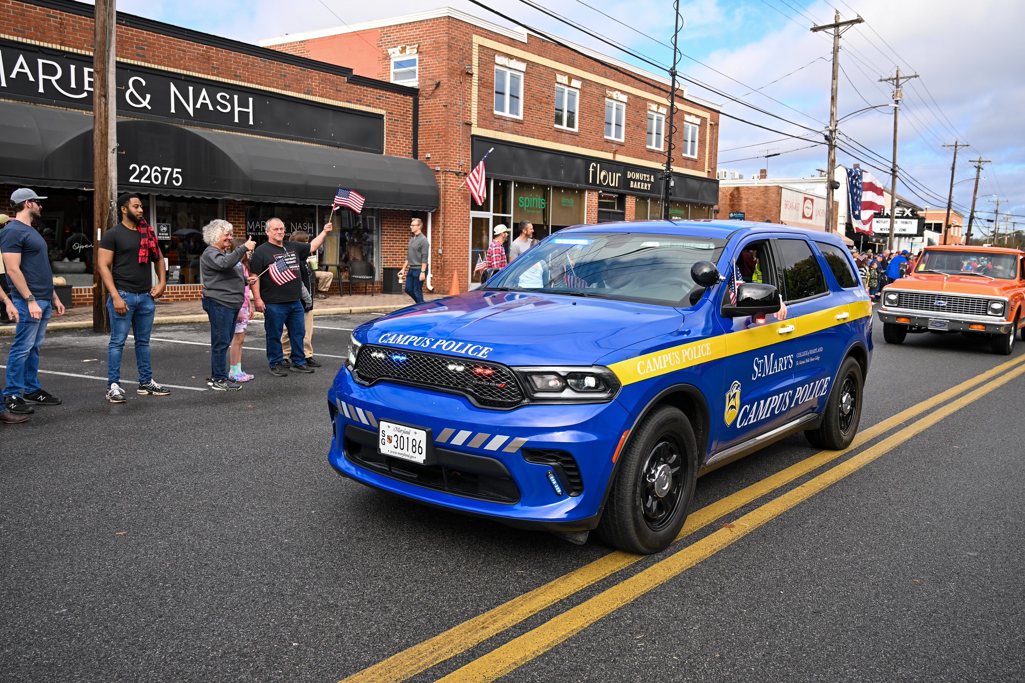 A blue campus police SUV drives down a street during a parade, with people holding American flags standing on the sidewalk in front of storefronts.