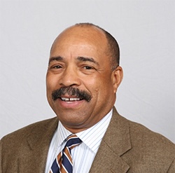 Man in a brown suit jacket, white dress shirt, and striped tie, standing and smiling in front of a plain light background.