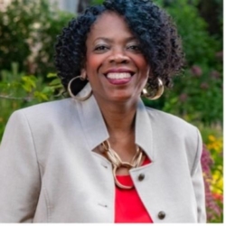 Woman with curly hair wearing a light beige blazer, red top, and gold jewelry, smiling outdoors with greenery in the background.