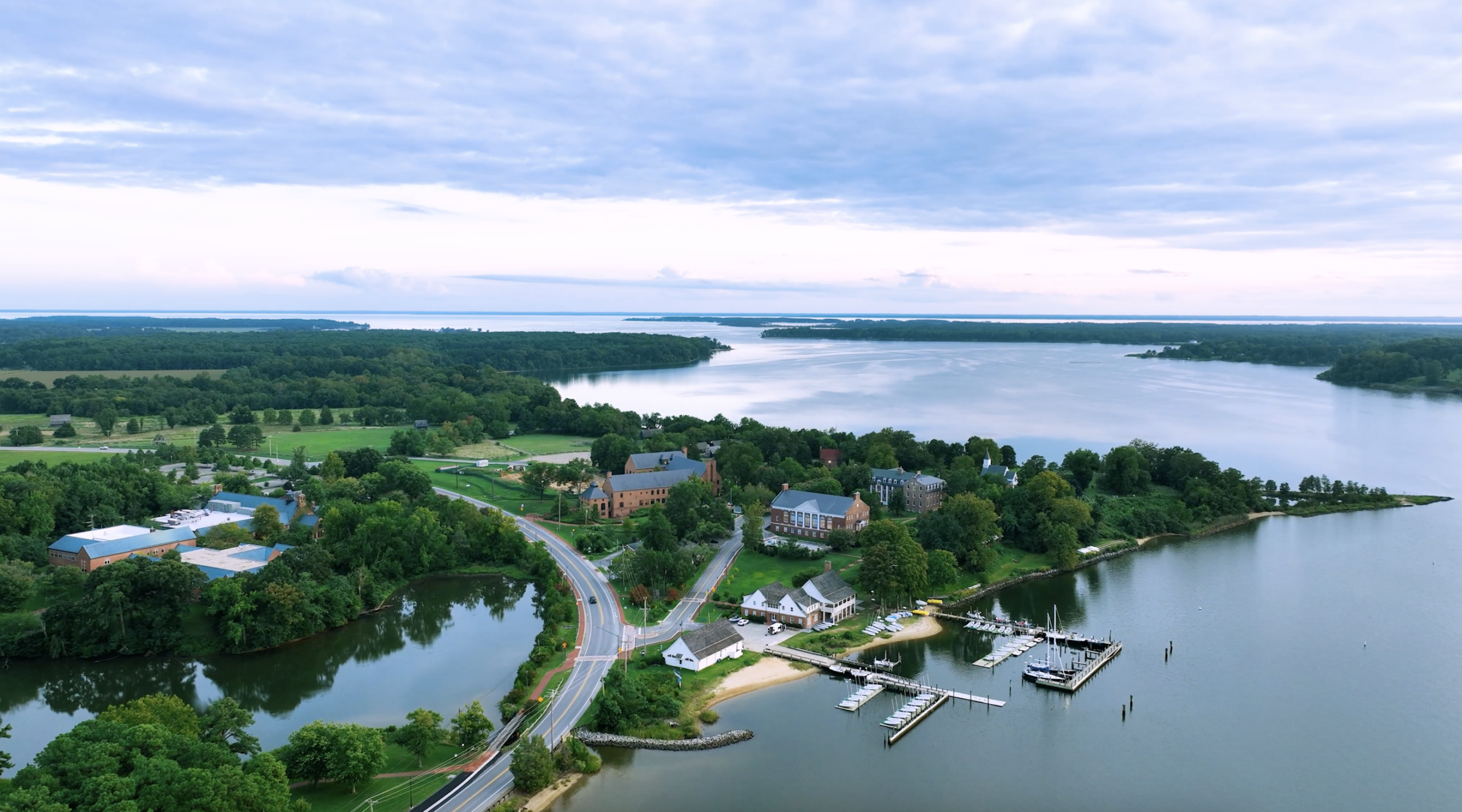 Aerial view of a waterfront community with houses, roads, docks, boats, and trees near a large body of water under a cloudy sky.