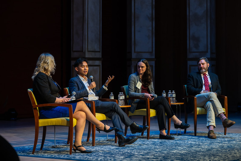 Four people sit in chairs on a stage having a panel discussion; one man speaks into a microphone while the others listen.