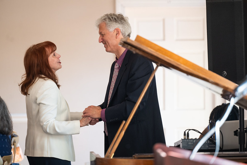 A woman and a man stand facing each other, holding hands and talking, with a grand piano in the foreground.