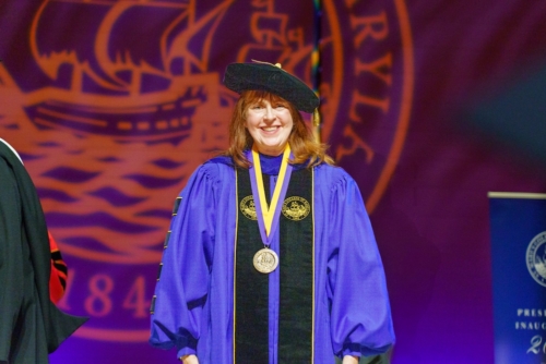 A woman in academic regalia with a medal around her neck stands in front of a purple backdrop featuring a ship and university seal."