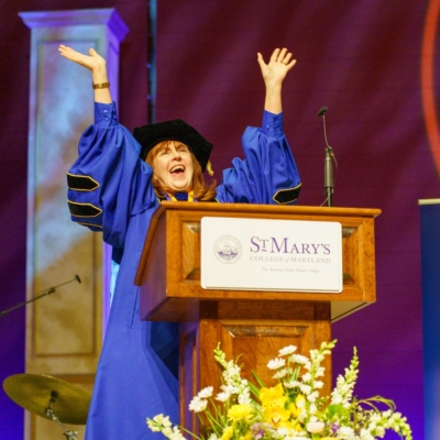 A person in academic regalia stands at a podium labeled "St. Mary's College of Maryland," raising their hands and speaking enthusiastically at a formal event.