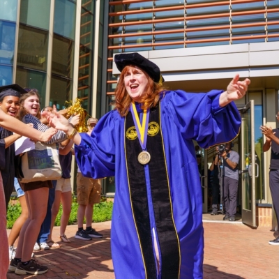 A person in a blue academic gown and cap stands outside a building, smiling and holding out their arms, as people nearby clap and cheer.