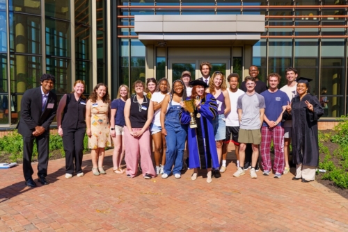 A group of people, some in academic regalia, pose outside a modern building. Two hold a trophy and flash peace signs. It is sunny and everyone is smiling."