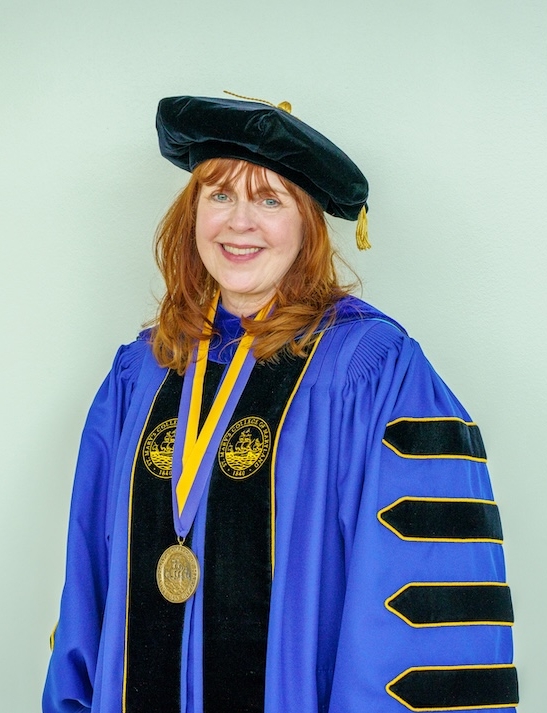 A woman in academic regalia with a black cap, blue gown with black and gold trim, and a medal, standing against a light background.