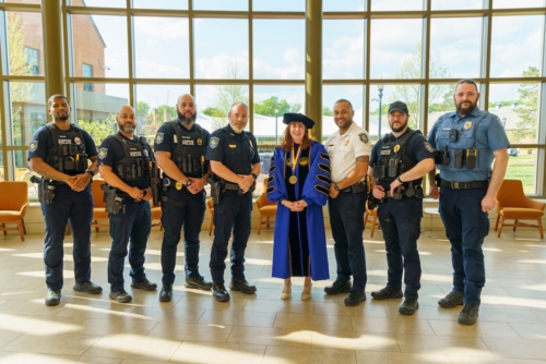 Eight people, seven in police uniforms and one in academic regalia, stand in a line inside a bright building with large windows and chairs in the background."
