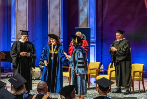 A group of people in academic regalia stand on a stage during a graduation ceremony, smiling and clapping."