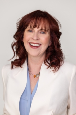 A woman with curly red hair and bangs, wearing a white blazer over a light blue top, smiles at the camera against a plain light background.