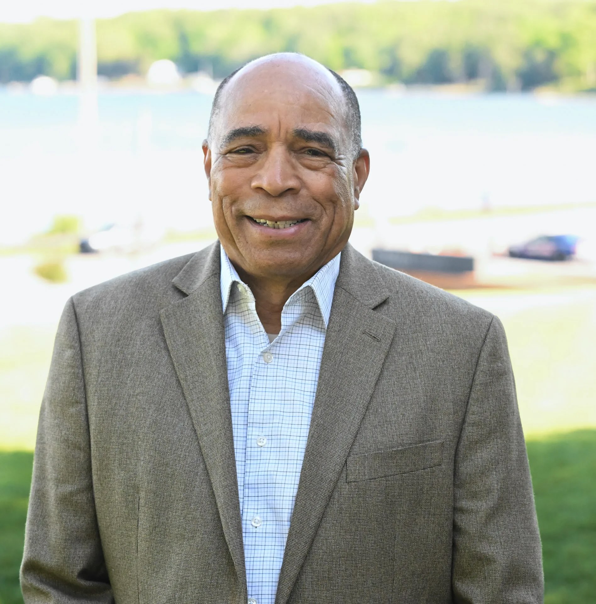 A man in a brown blazer and checked shirt stands outside on a sunny day, with greenery and water in the blurred background.