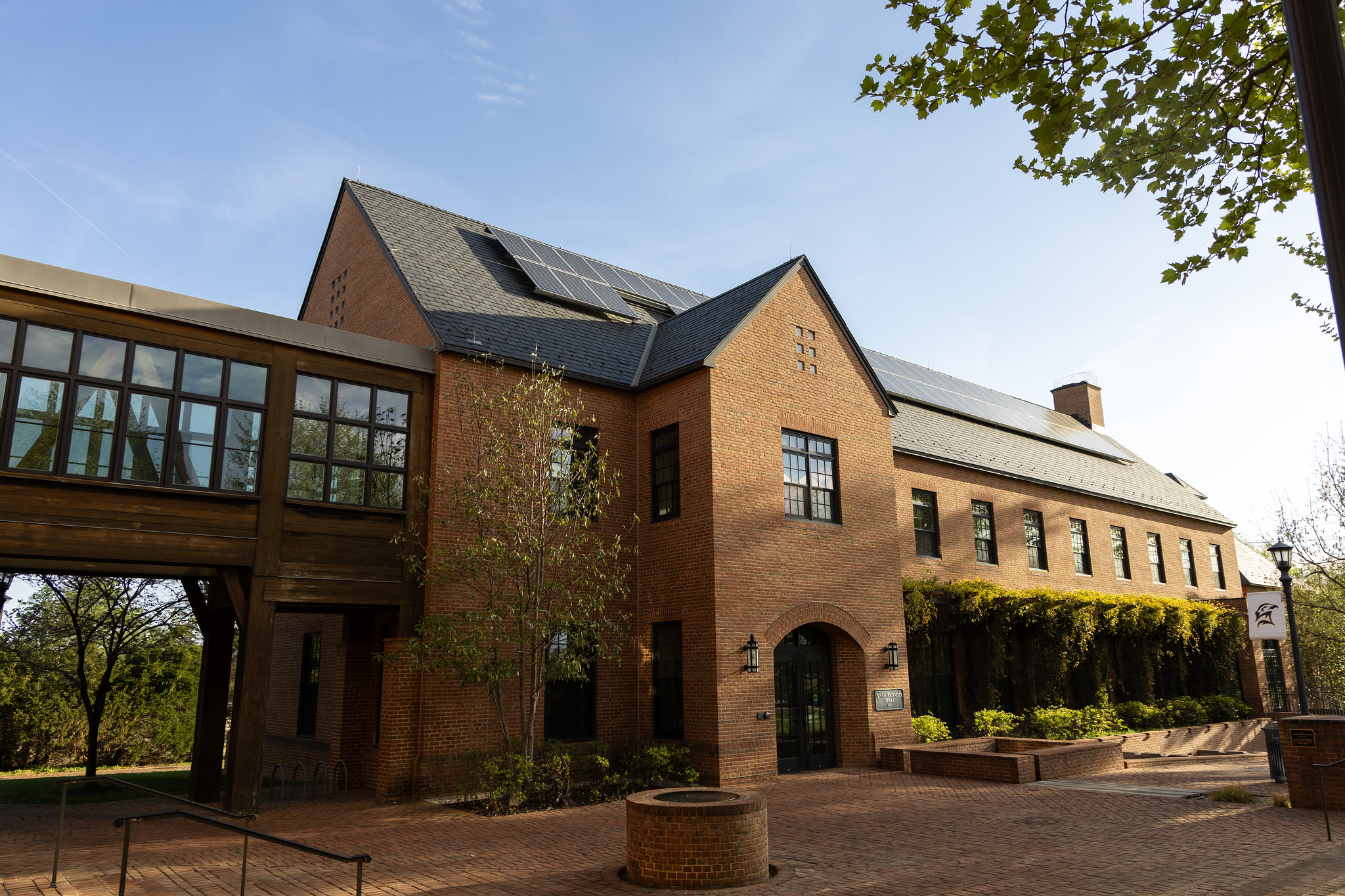 A brick building with a glass walkway and solar panels on the roof, surrounded by trees and a brick courtyard under a clear sky.