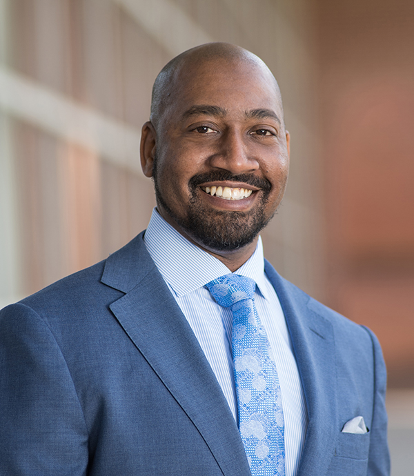 A man in a blue suit and tie smiles at the camera while standing in front of a blurred building background.