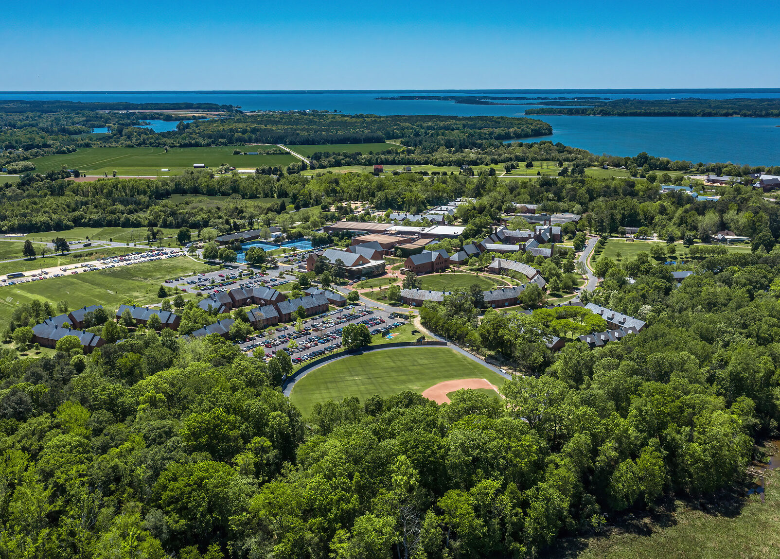 Aerial view of a campus with multiple buildings, roads, parking lots, a baseball field, and surrounding trees near a large body of water.