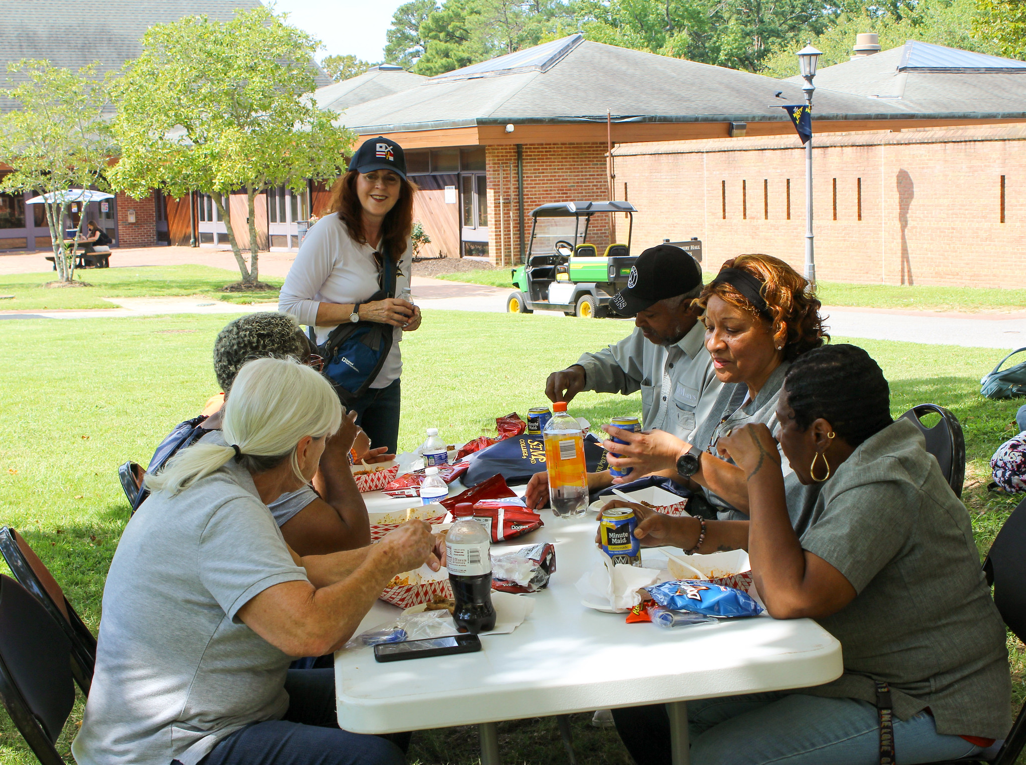 A group of people sitting at a picnic table eating lunch outdoors, with a woman standing nearby.
