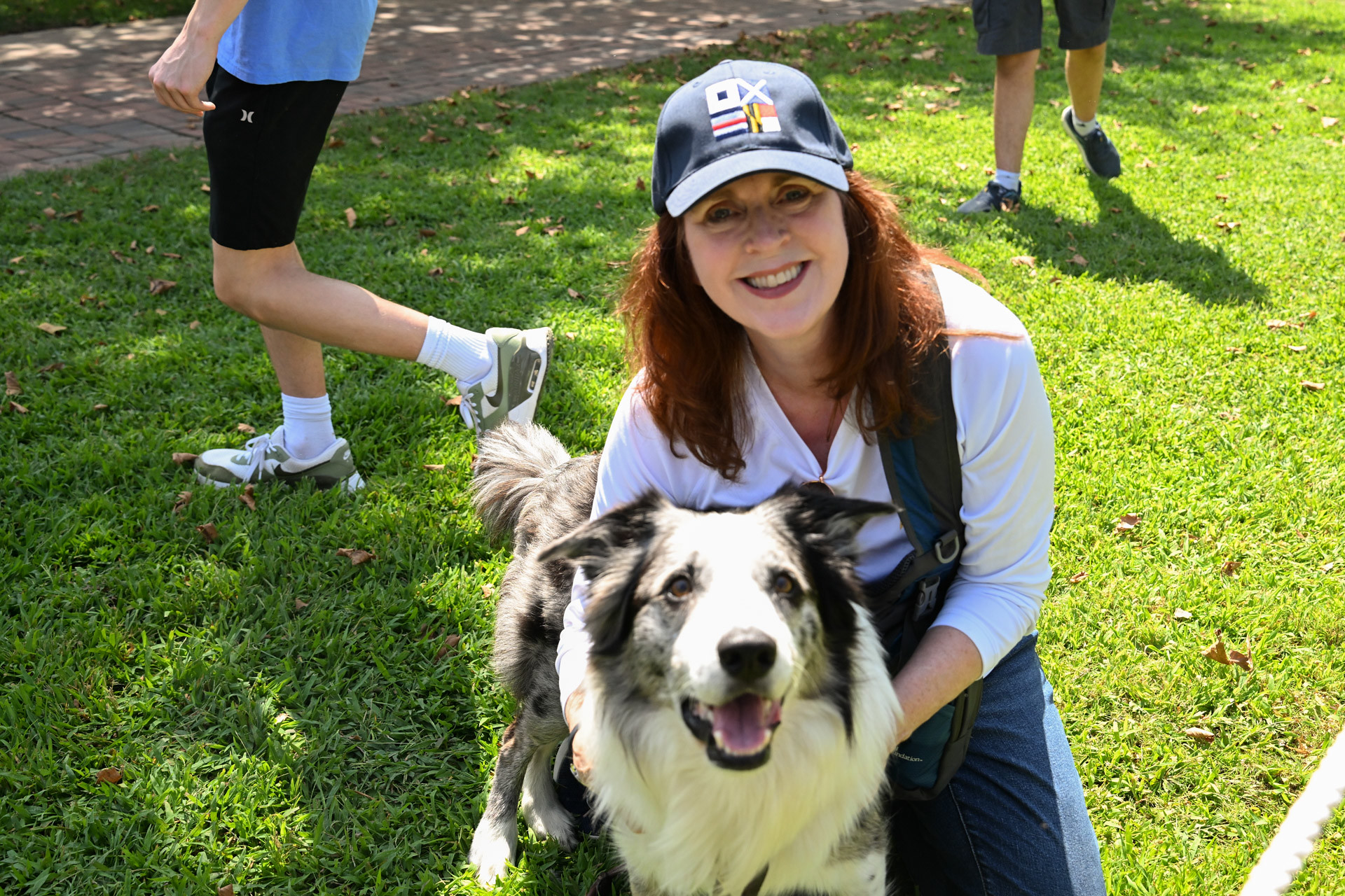 A woman in a baseball cap smiles while kneeling on the grass with a border collie.