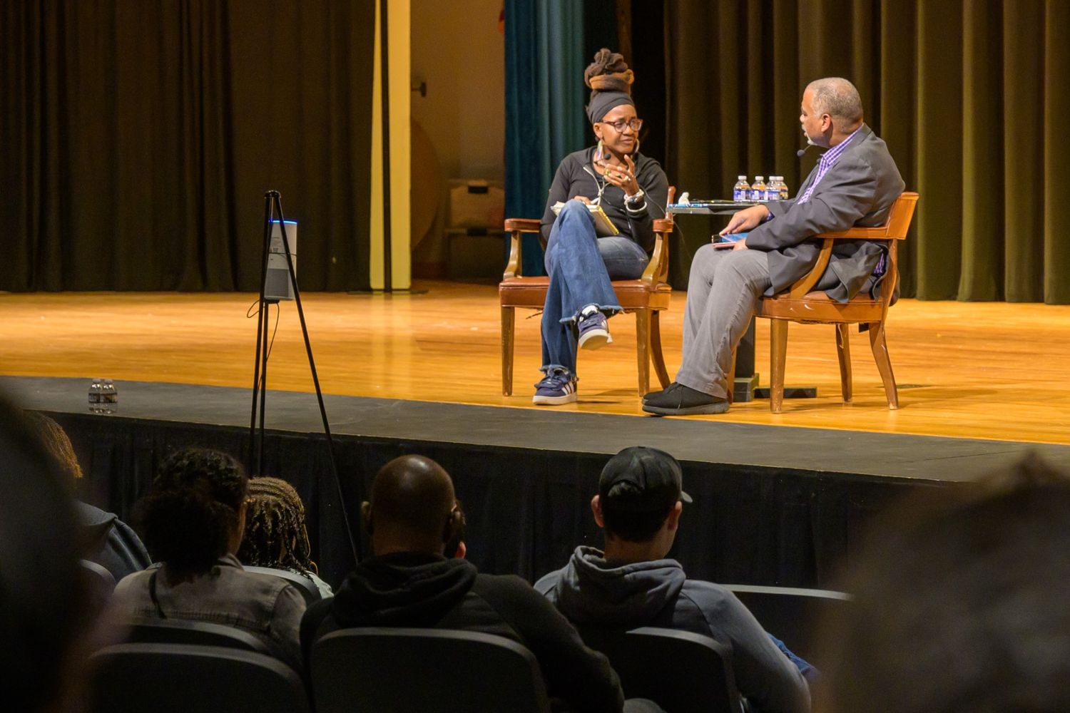 54068863282_d98f9a19be_o (1) Two people sit on stage having a discussion while an audience watches from the seats in front of them. There are bottled waters on the table between the speakers.