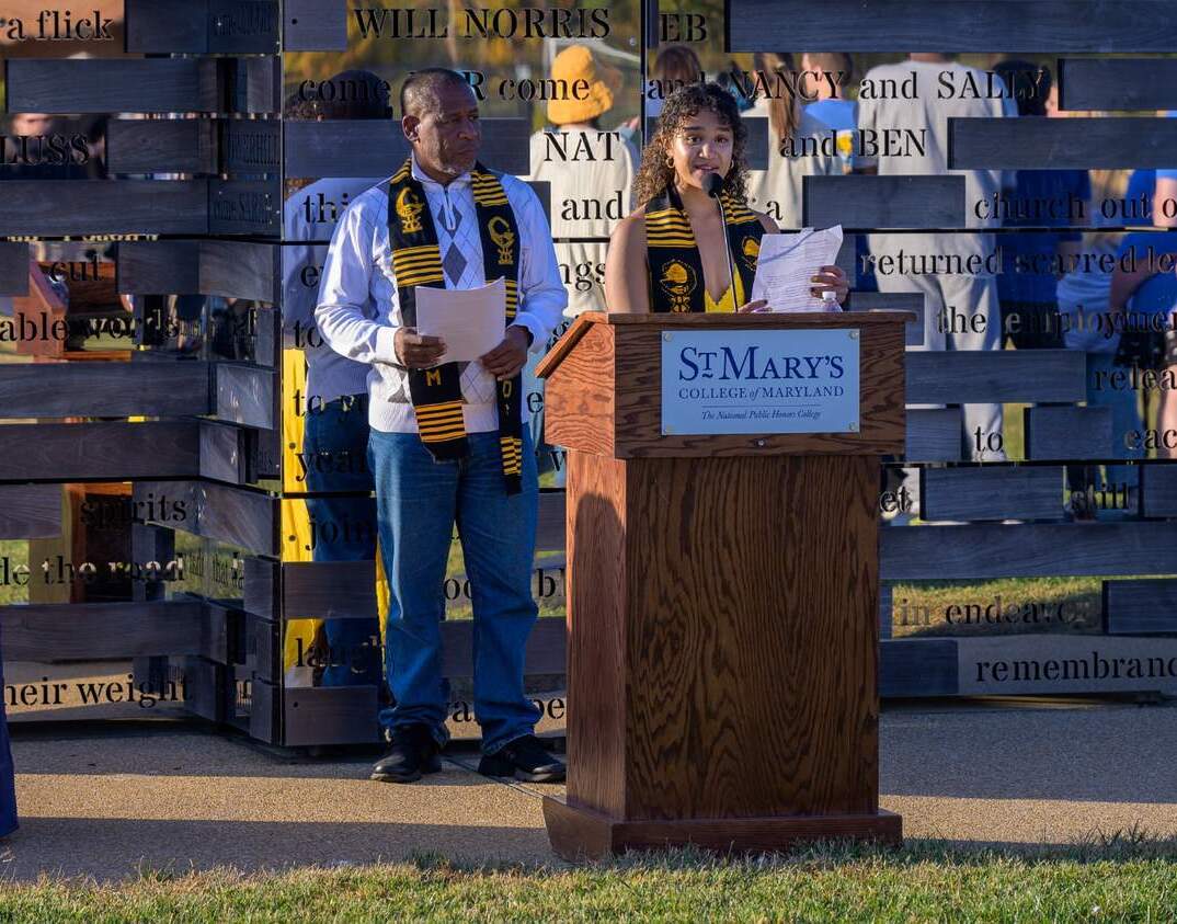 54123321476_a063f1579b_o (1) Two people stand at a wooden podium labeled "St. Mary's College of Maryland," speaking at an outdoor event; both wear academic stoles.