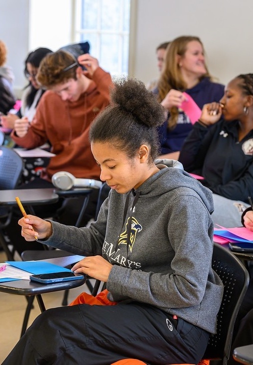 54277941241_bd40377460_c A student writes on a notepad at a desk in a classroom, while other students sit and interact in the background.