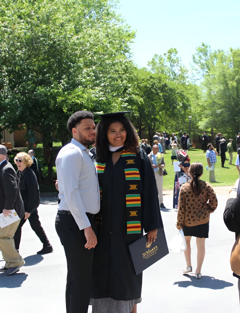 54516837006_380fabca76_c (1) A graduate in cap and gown poses with a man outdoors after a ceremony, surrounded by other people on a sunny day.