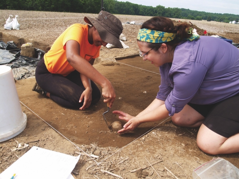 Two people participate in an archaeological dig, carefully uncovering an object from the soil within a marked excavation site outdoors.