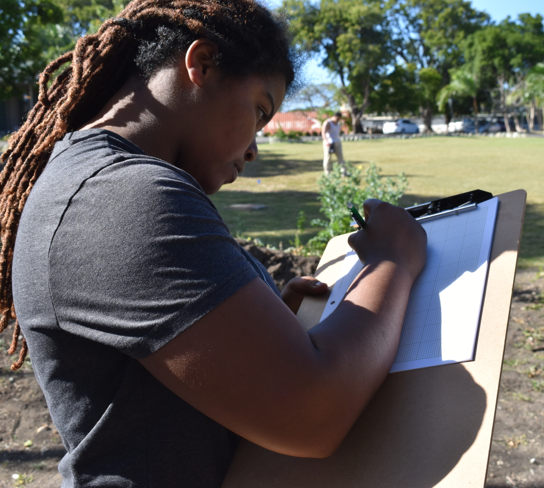 Person holding a clipboard and writing on a chart outdoors in a grassy area with trees and people in the background.
