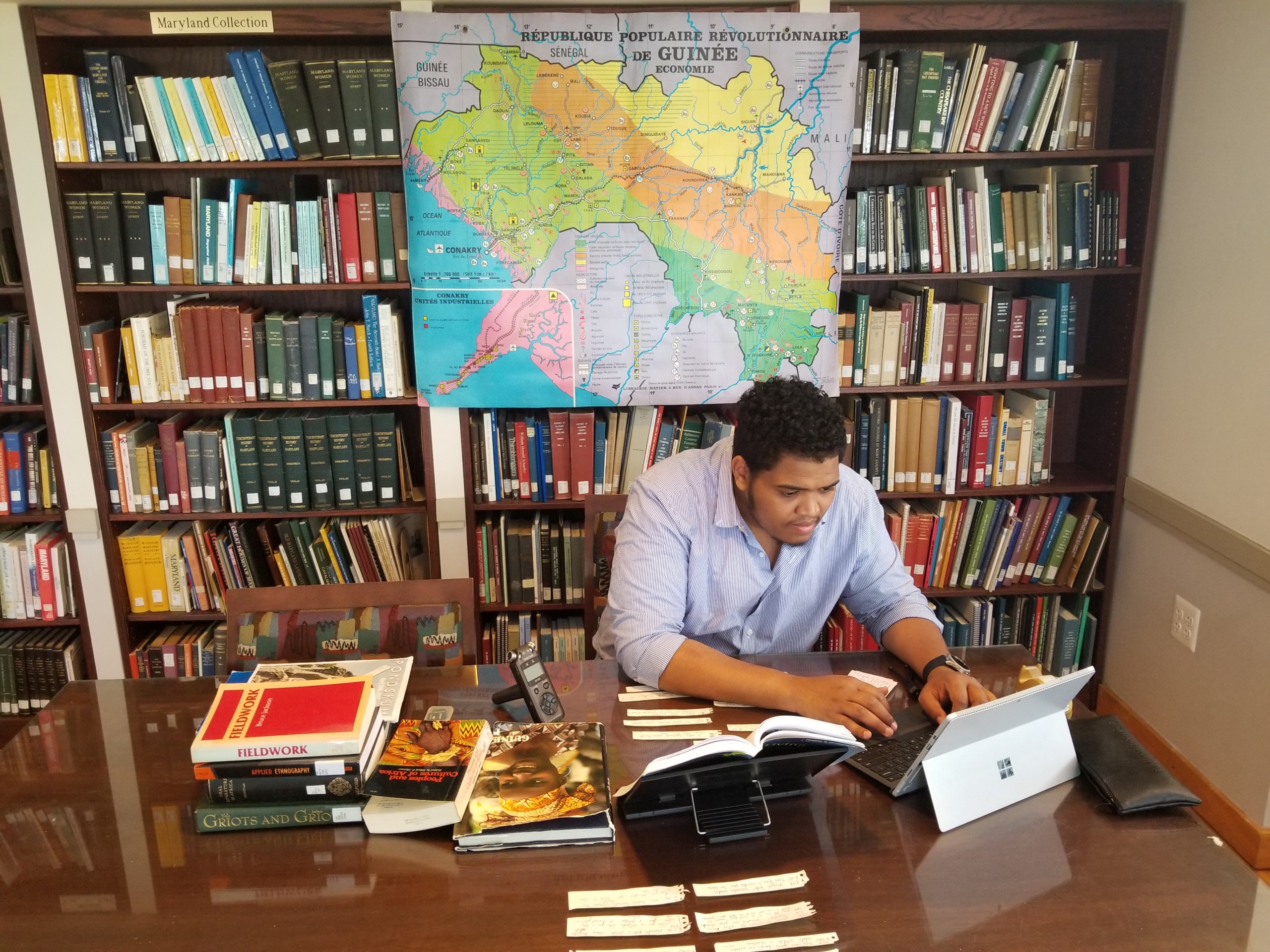Zeid Chleuh at work preparing to head to Guinea A person sits at a desk in a library, working on a laptop surrounded by books and papers, with a map of Guinea displayed on the bookshelf behind them.