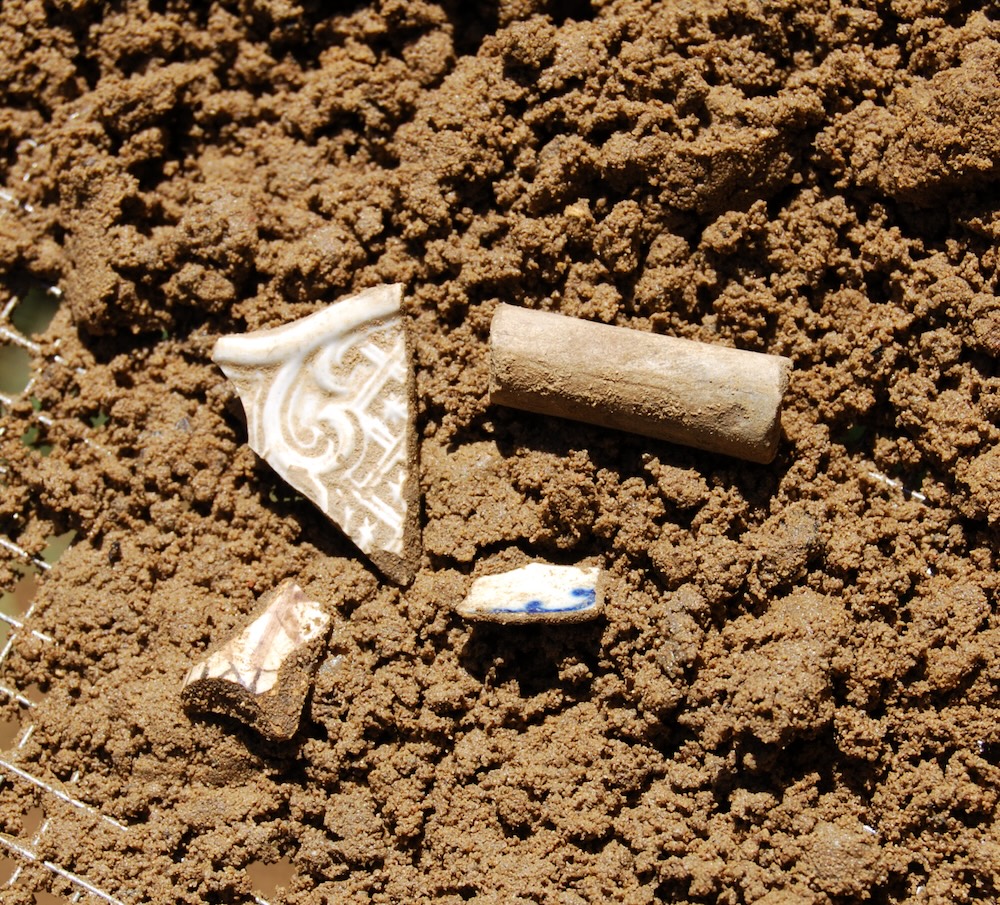 Four small pottery and ceramic fragments partially buried in brown soil, including a decorated pottery shard, a cylindrical object, and two smaller pieces.