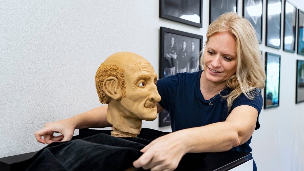 A woman unveils a clay bust of a bald man with prominent ears and mustache, next to a white wall with framed photos in the background.