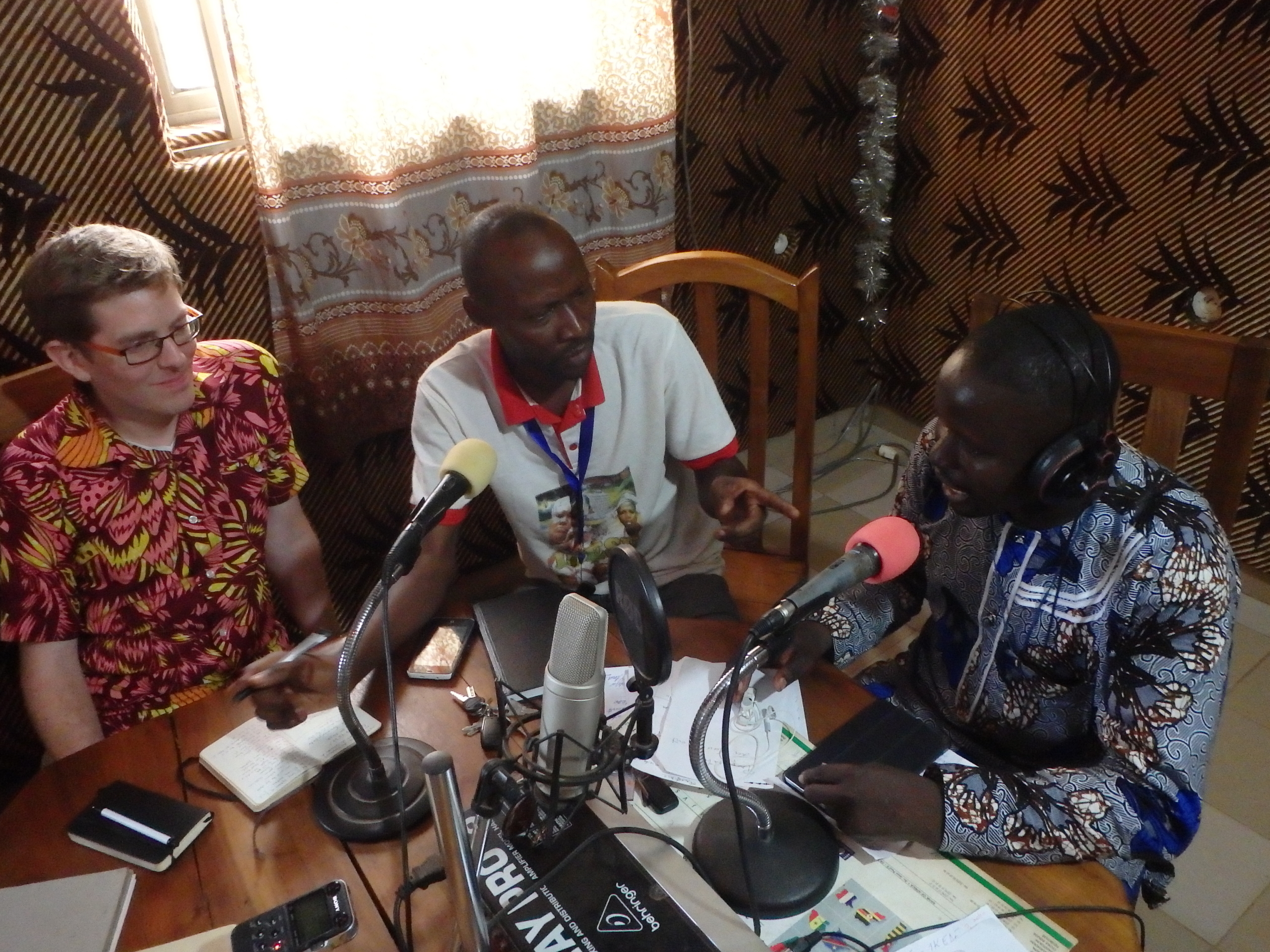 Three men sit at a table in a radio studio, speaking into microphones, with notebooks and papers in front of them.